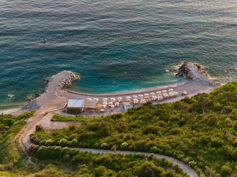 Luftaufnahme eines abgelegenen Strandes mit Liegestühlen und Sonnenschirmen neben grün bewachsenem Hügel.