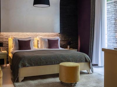 Modernes Hotelzimmer mit großem Bett, grauem Bettüberwurf und gelbem Hocker vor einem Fenster mit Vorhängen.