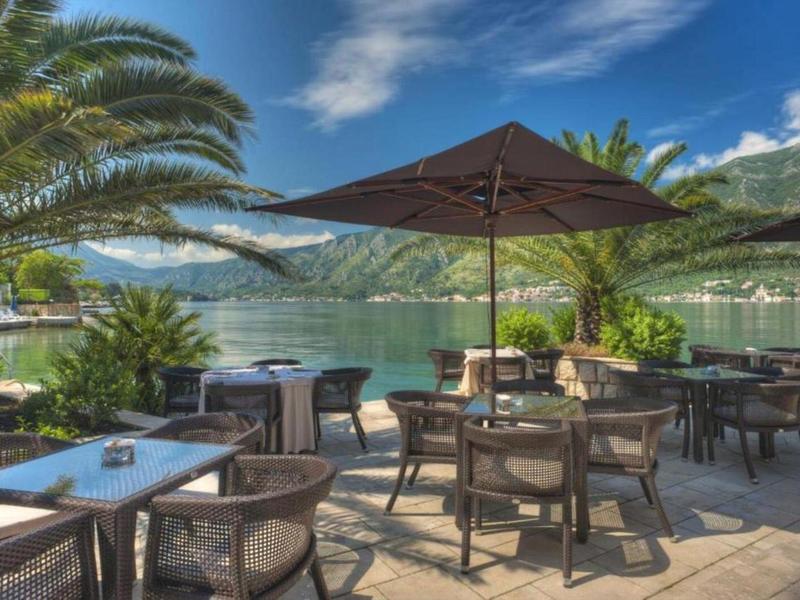 Terrasse mit Tischen und Stühlen unter Sonnenschirmen am See mit Bergen im Hintergrund