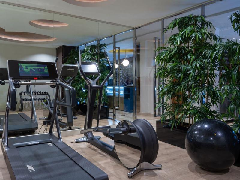 Fitnessraum mit Laufband, Rudergerät, Gymnastikball und großen Pflanzen in modernem Ambiente.