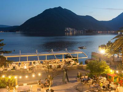 Abendliche Strandbar mit Tischen und Stühlen, beleuchtet, vor ruhigem Meer und Bergen.