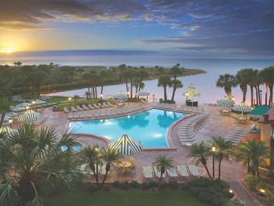 Poolbereich eines Hotels mit Palmen und Sonnenuntergang über dem Meer im Hintergrund.