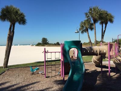 Kinderspielplatz mit Rutsche, Klettergerüst, Schaukeln und Palmen bei klarem Himmel.
