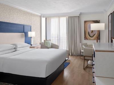 Helles Hotelzimmer mit großem Bett, Schreibtisch, Fernseher und Bodenvorhängen.