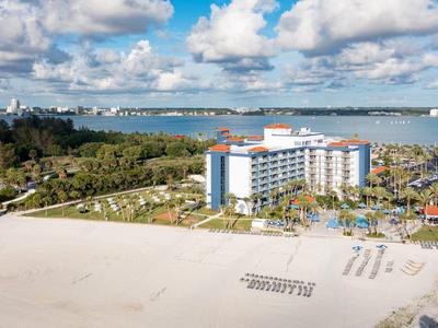 Großer Sandstrand, weißes Hotelgebäude, grüne Vegetation und blauer Himmel mit Wolken über einer Wasserfläche.