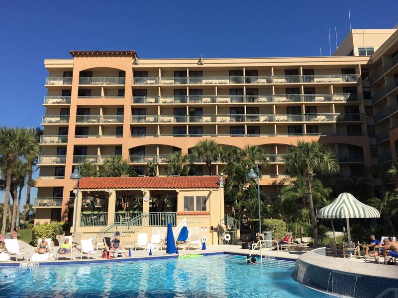 Mehrstöckiges Hotel mit Balkon, Palmen und großem Außenpool unter klarem blauem Himmel.