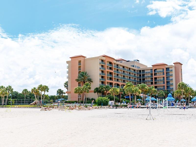 Ein großes Hotelgebäude am Strand mit Palmen, blauem Himmel und Sand im Vordergrund.