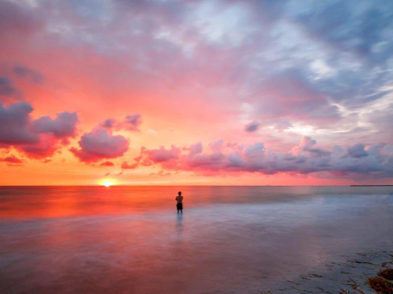 Farbenfroher Sonnenuntergang über ruhigem Meer mit Person im Wasser und Wolken am Himmel.