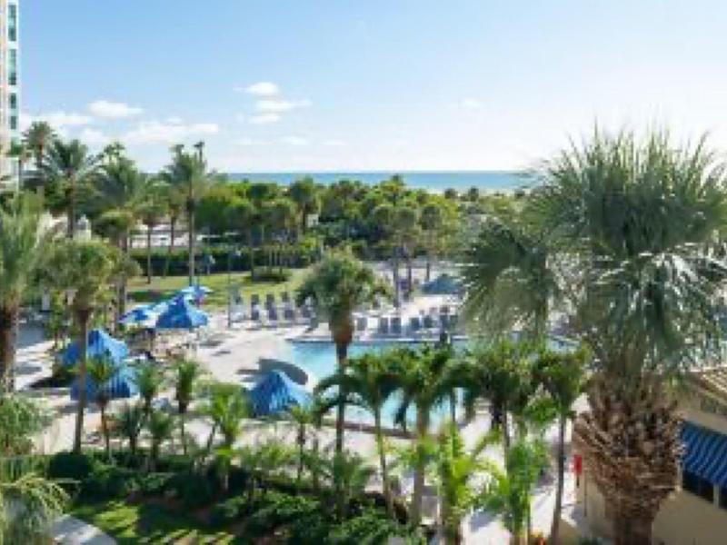 Blick auf einen Hotelpool mit blauen Sonnenschirmen, Palmen und Meer im Hintergrund.