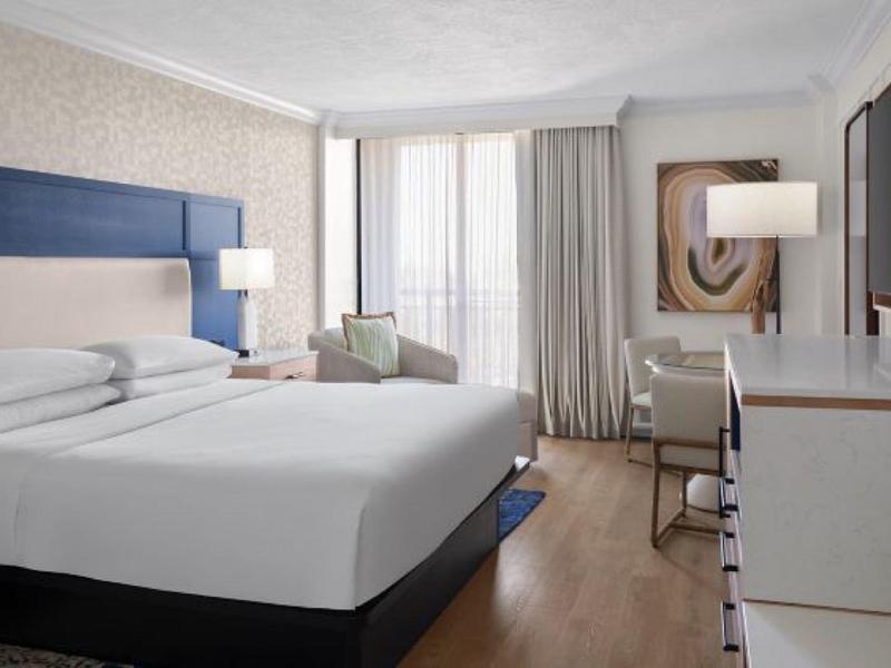 Helles Hotelzimmer mit großem Bett, Schreibtisch, Fernseher und Bodenvorhängen.