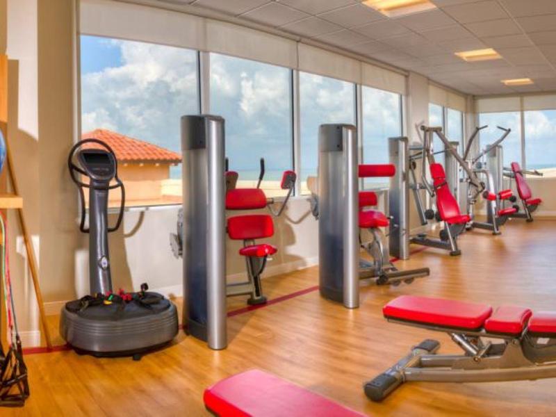 Ein heller Fitnessraum mit großen Fenstern, roten Trainingsbänken und verschiedenen Geräten auf Holzboden.