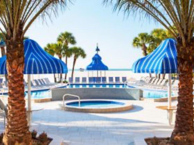 Poolbereich mit blau-weiß gestreiften Sonnenschirmen, Liegestühlen und Palmen bei sonnigem Wetter.