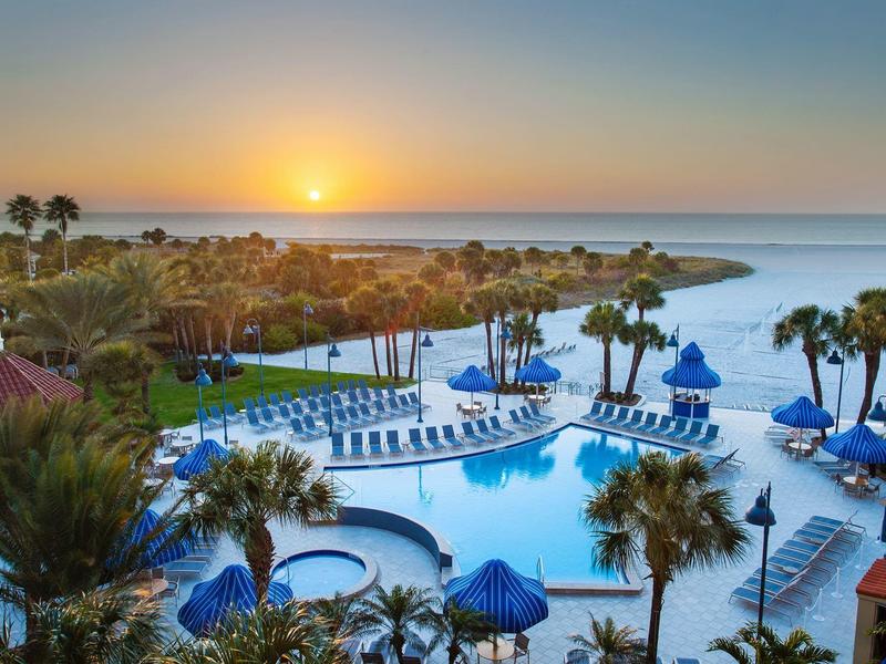 Strandhotel mit Pool, Palmen und Sonnenuntergang über ruhigem Meer.