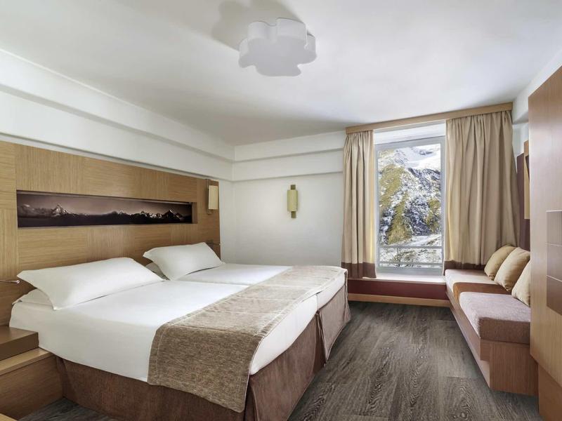 Světle zařízený hotelový pokoj se dvěma postelemi, lavicí u okna a výhledem na hory.