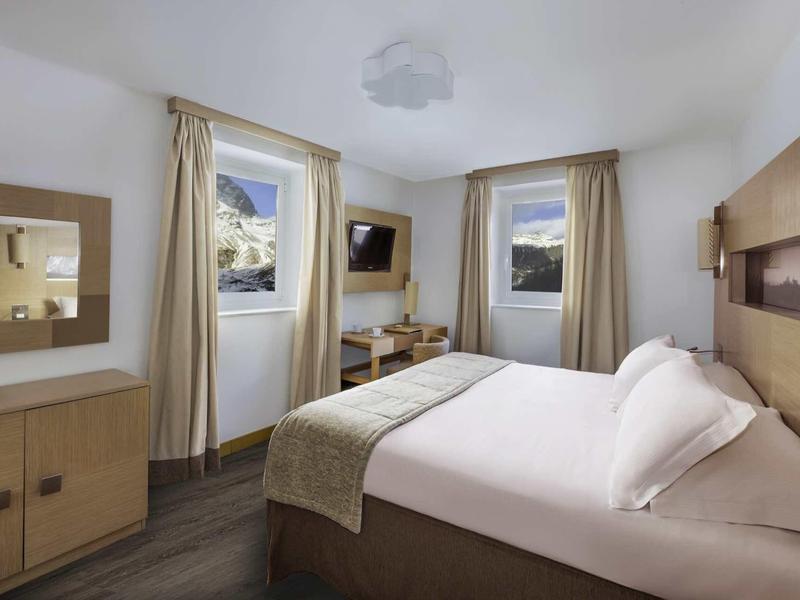 Světý hotelový pokoj s manželskou postelí, dvěma okny, televizí, zrcadlem a dřevěným nábytkem.