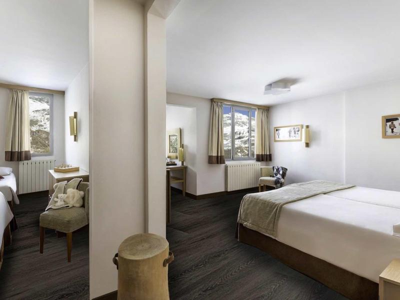 Jasně zařízený hotelový pokoj se dvěma jednolůžky a manželskou postelí, moderním nábytkem a velkými okny.