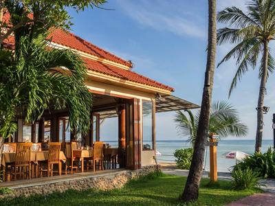 Offenes Strandrestaurant mit Holzmöbeln, Palmen und Blick auf das ruhige, blaue Meer.
