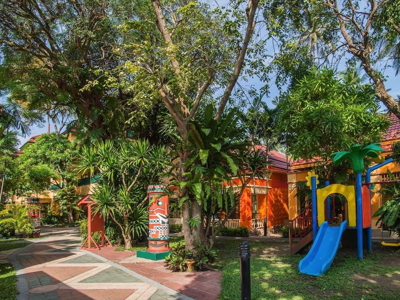 Bunter Spielplatz mit Rutsche, tropischen Bäumen und bunten Gebäuden im Sonnenschein.