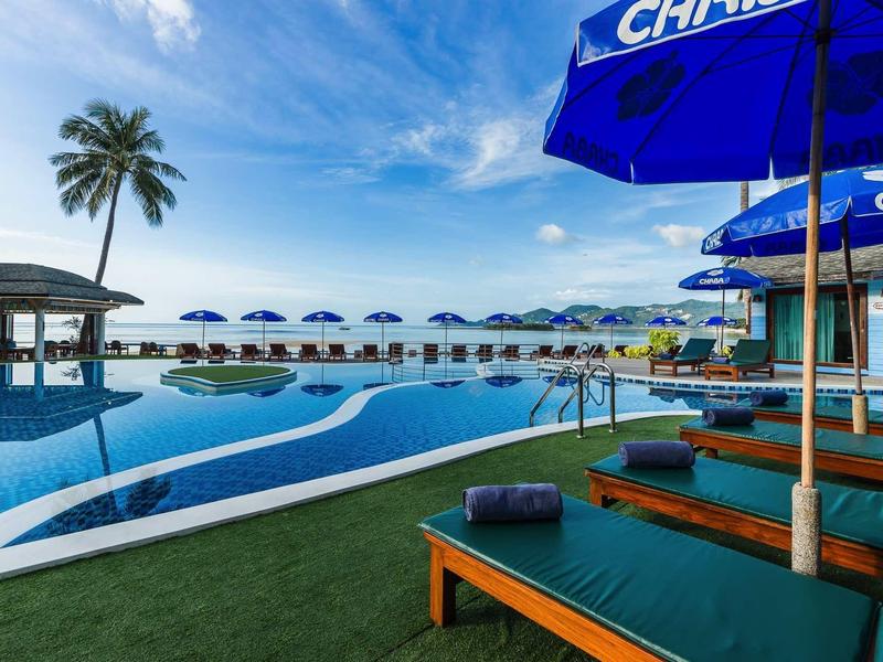 Strandresort mit Pool, blauen Sonnenschirmen, Liegen und Palmen unter blauem Himmel.