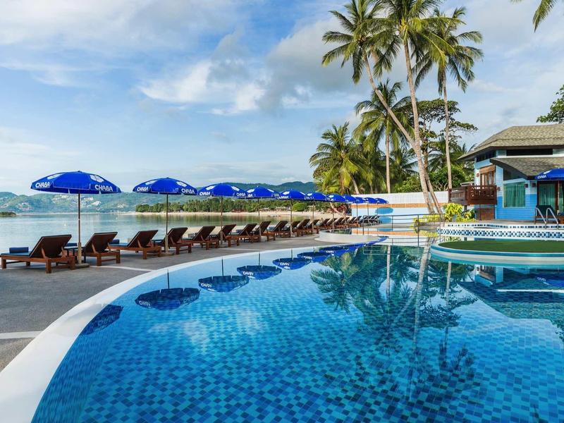 Schwimmbecken mit blauen Kacheln, Liegestühlen, blauen Sonnenschirmen und Palmen am Meer.