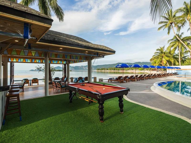 Freier Billardtisch auf grünem Kunstrasen neben Pool und Strand mit Palmen und Sonnenschirmen.