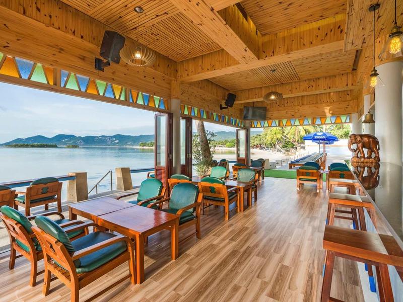 Offene Holzterrasse mit Holztischen, bunten Sitzpolstern und Blick auf Meer und Berge.