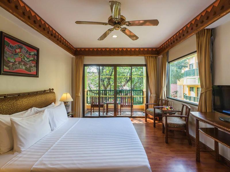 Helles Schlafzimmer mit großem Bett, Deckenventilator, Fernseher, Holzfußboden und Balkon mit Blick ins Grüne.