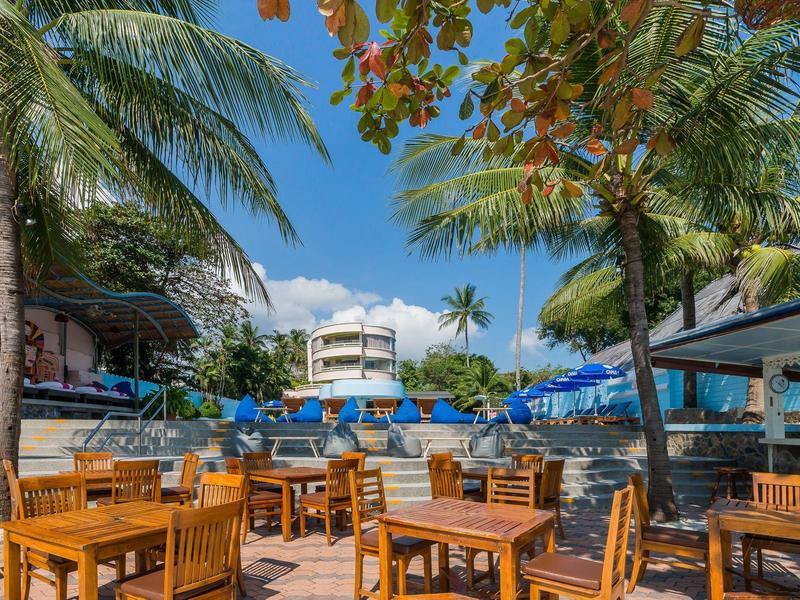 Ristorante all'aperto con tavoli in legno, palme e cielo azzurro vicino a una piscina dell'hotel.