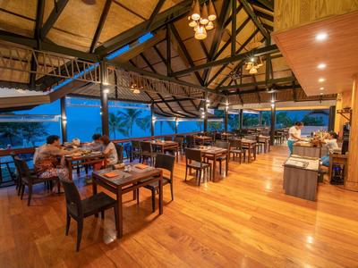 Offenes Restaurant mit Holzboden und Dach, zahlreichen Tischen, Stühlen und Blick auf den Abendhimmel.