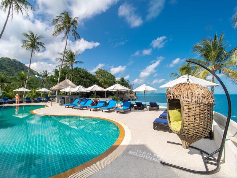 Schwimmbad mit blauem Wasser, Liegestühlen, Sonnenschirmen und Palmen vor blauem Himmel und Bergen.