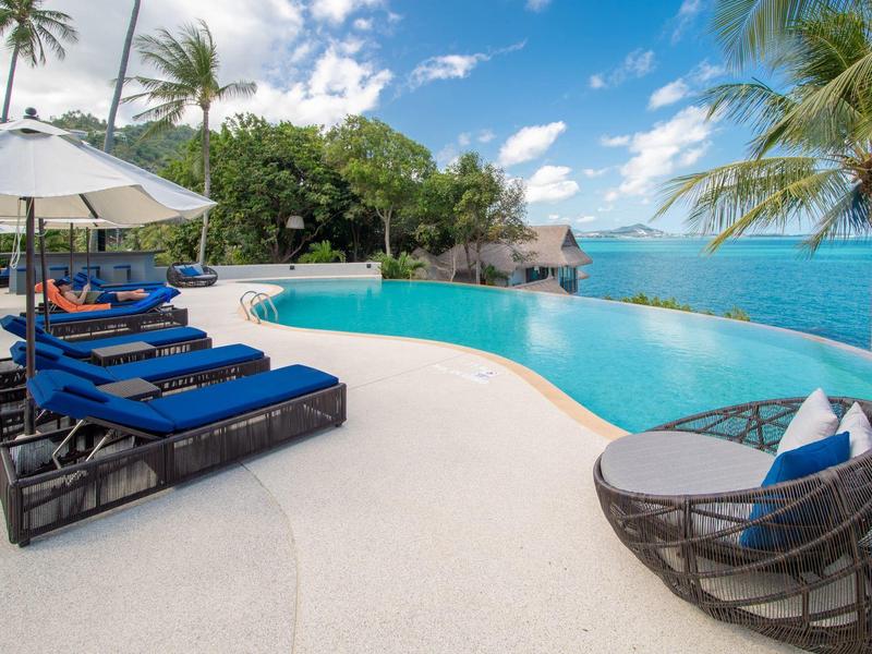 Infinity-Pool mit blauen Liegen, Palmen und Meerblick unter blau-weißem Himmel.