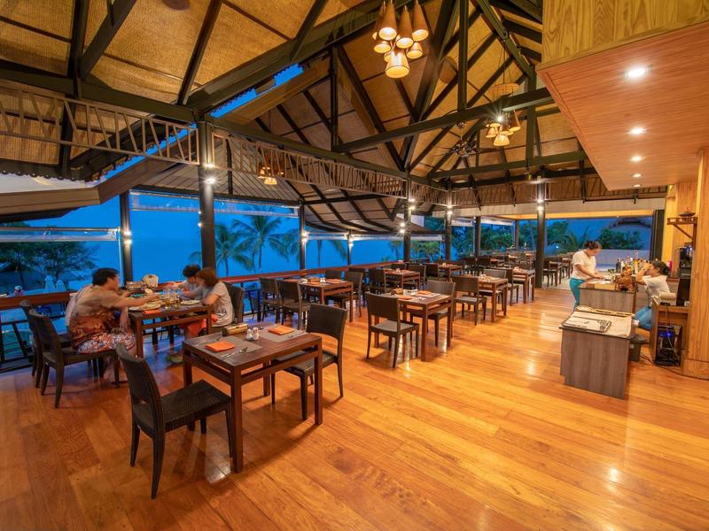 Offenes Restaurant mit Holzboden und Dach, zahlreichen Tischen, Stühlen und Blick auf den Abendhimmel.