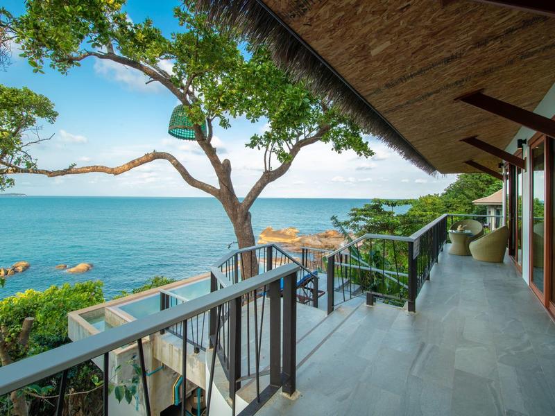 Balkon mit Fliesenboden, Holzgeländer, Blick auf Baum und blaues Meer unter klarem Himmel.