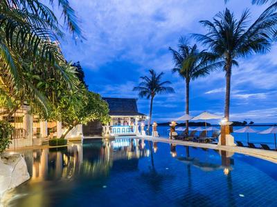 Luxuriöser Pool mit Palmen am Abend, blaues Wasser, beleuchtete Gebäude und bewölkter Himmel.