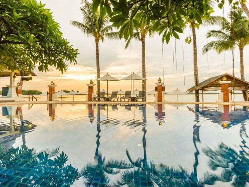 Una piscina de hotel con palmeras y sombrillas en la playa al atardecer.