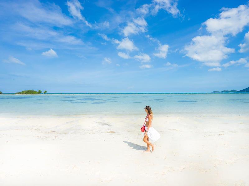 Mujer con vestido rojo camina por la playa de arena blanca con cielo azul y mar al fondo.