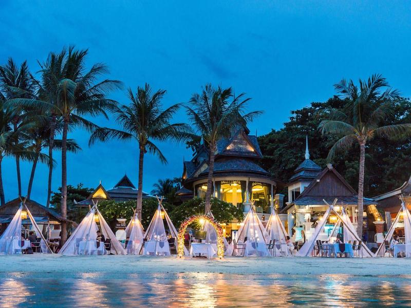 Resort de playa al anochecer con tipis iluminados, palmeras y piscina en primer plano.