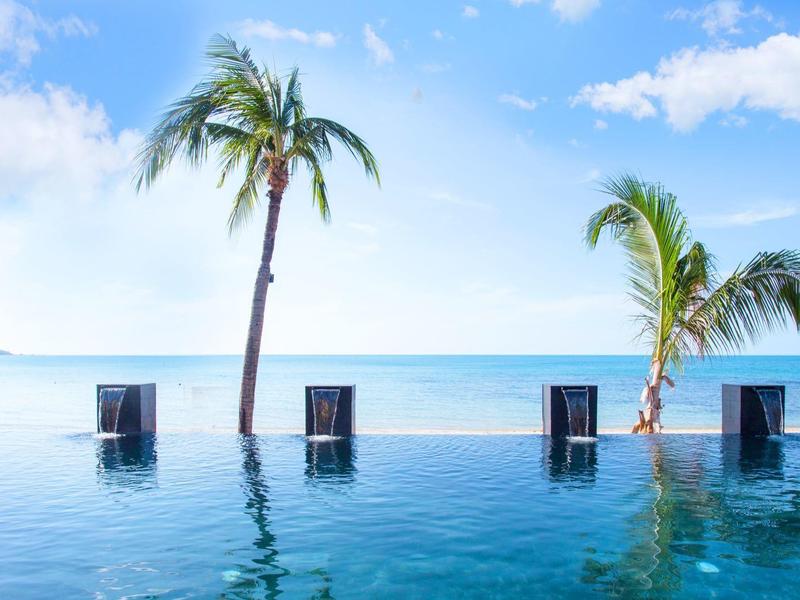 Piscina a sfioro con quattro sedie e palme, vista sul mare calmo e cielo azzurro.