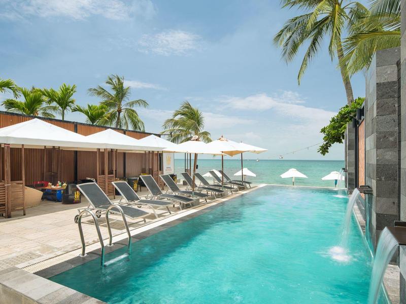 Piscina moderna con lettini e ombrelloni accanto a palme tropicali e vista sul mare.