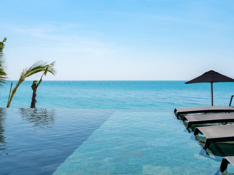 Piscina a sfioro accanto al mare con lettini e ombrellone sotto un cielo azzurro.