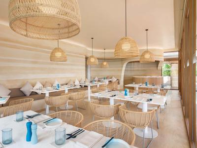 Helles Restaurant mit Holzstühlen, weißen Tischen und geflochtenen Lampen in warmem Beige.