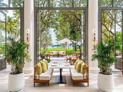 Eleganter Lounge-Bereich mit weißem Boden, Holzsofas und grünen Pflanzen, Blick auf Garten mit Pool.