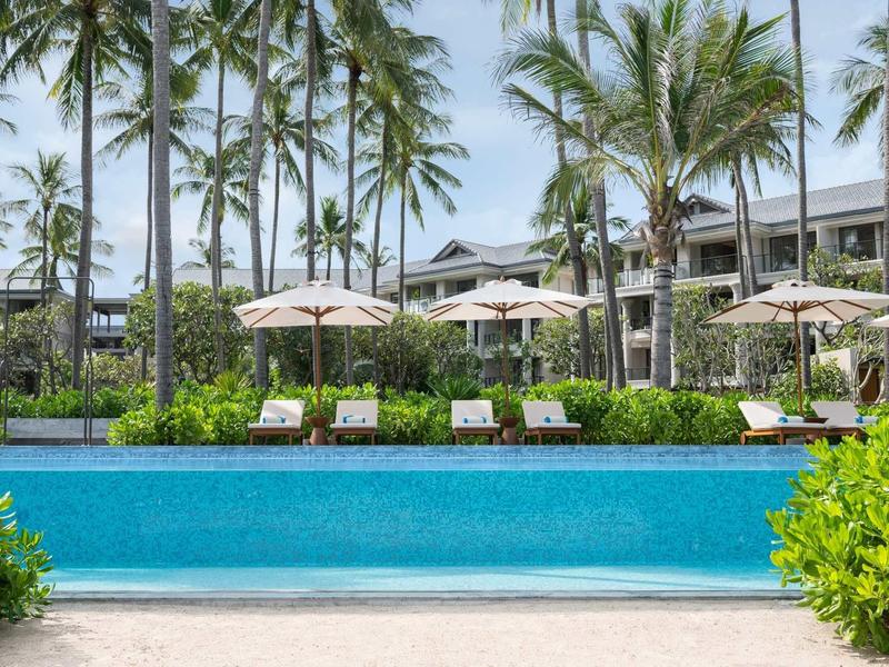 Moderner Poolbereich mit Liegestühlen und Sonnenschirmen umgeben von Palmen in einem Hotel.