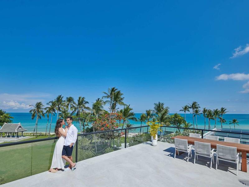 Paar in weißer Kleidung steht auf Terrasse mit Blick auf Palmen und türkisblaues Meer unter blauem Himmel.