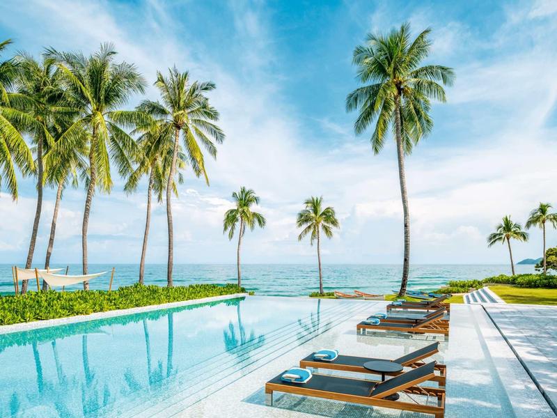 Infinity-Pool mit Liegestühlen, umgeben von hohen Palmen und Blick auf das blaue Meer unter blauem Himmel.