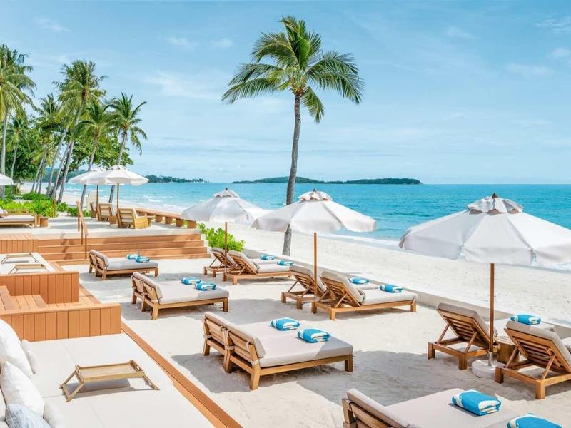 Strandterrasse mit Liegestühlen, Sonnenschirmen, Palmen und blauem Meer unter klarem Himmel.