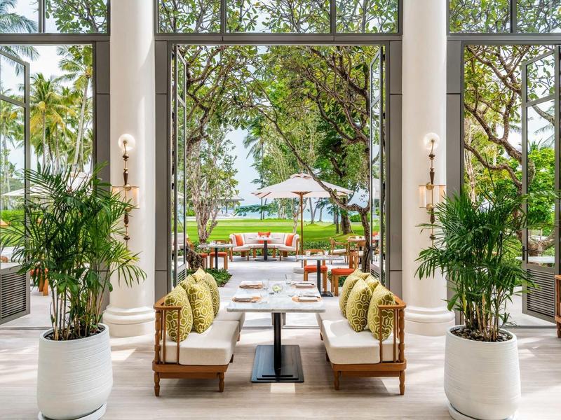 Eleganter Lounge-Bereich mit weißem Boden, Holzsofas und grünen Pflanzen, Blick auf Garten mit Pool.