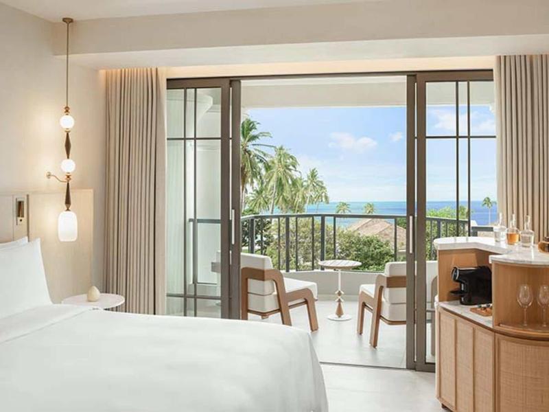 Helles Schlafzimmer mit weißem Bett, Holz Möbeln und Balkon mit Meerblick und Palmen.