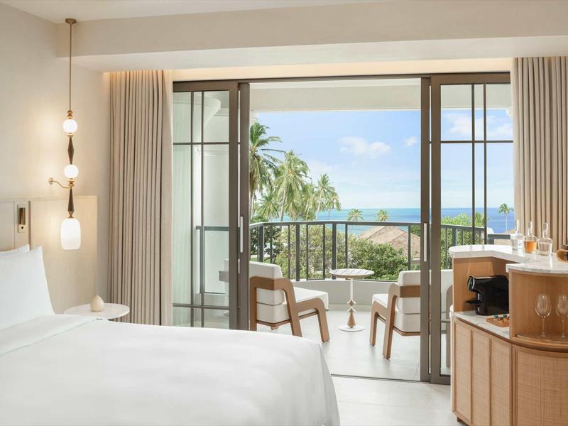 Helles Hotelzimmer mit weißem Bett und Balkon mit Meerblick.