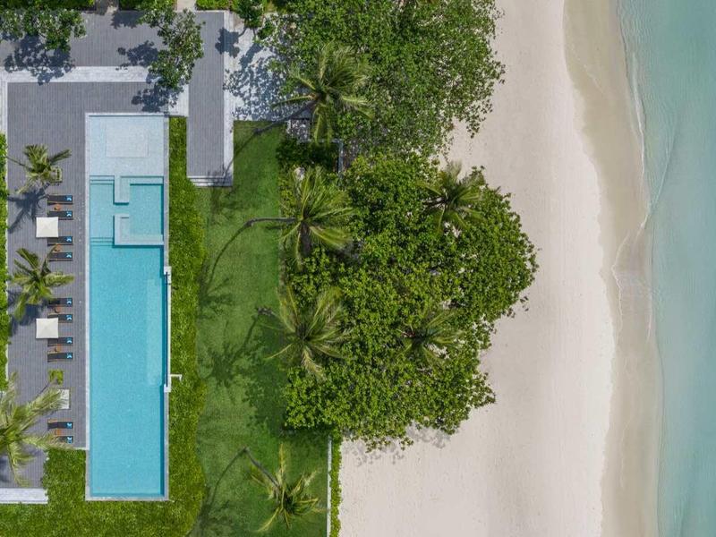 Luftaufnahme eines Pools neben grünem Garten und weißem Sandstrand am türkisfarbenen Meer.